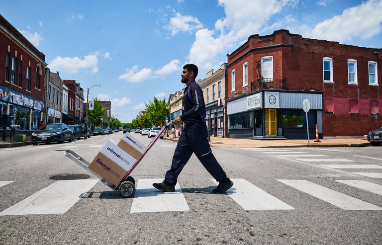 Man in business attire pulling a hand truck with boxes across a pedestrian crosswalk on a tree-lined downtown street